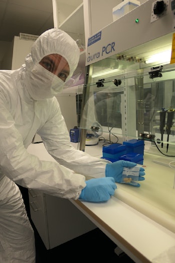 photo of scientist in a white biohazard suit with blue gloves standing at a counter next to a PCR ma...